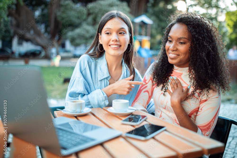 Two friends sit at a wooden table in a lively cafe, smiling and chatting while enjoying coffee. Laptops and phones are around them, creating a relaxed atmosphere. Two friends sit at a wooden table in a lively cafe, smiling and chatting while enjoying coffee. Laptops and phones are around them, creating a relaxed atmosphere.