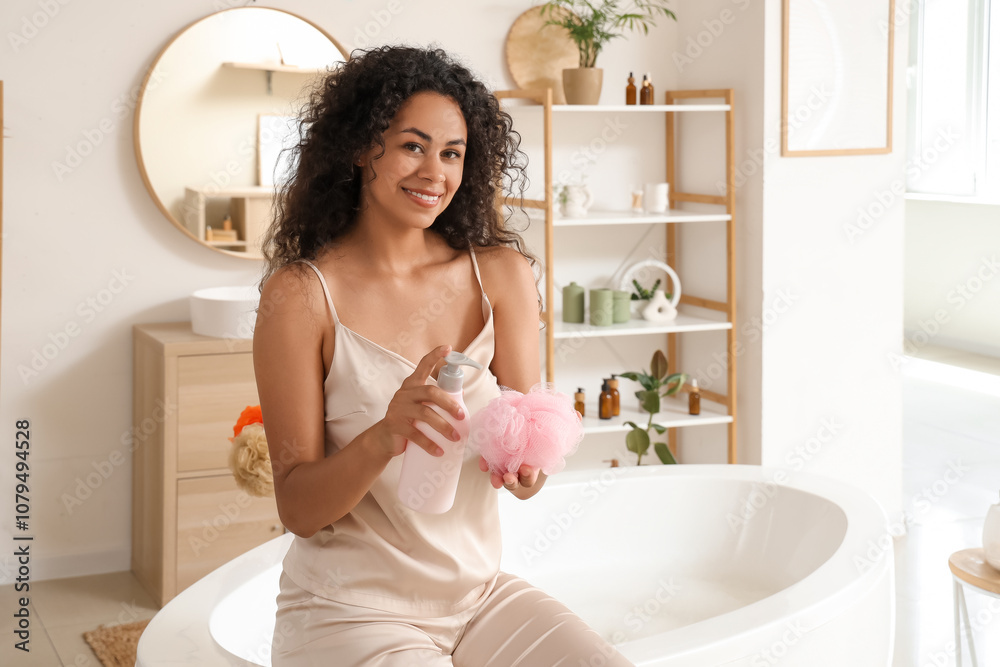 Beautiful young happy African-American woman with bottle of shower gel and bath sponge sitting at home Beautiful young happy African-American woman with bottle of shower gel and bath sponge sitting at home