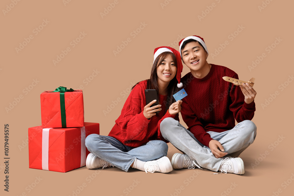 Beautiful young Asian couple in Santa hats with credit card, wooden airplane, mobile phone and gift boxes sitting on brown background. Christmas celebration Beautiful young Asian couple in Santa hats with credit card, wooden airplane, mobile phone and gift boxes sitting on brown background. Christmas celebration