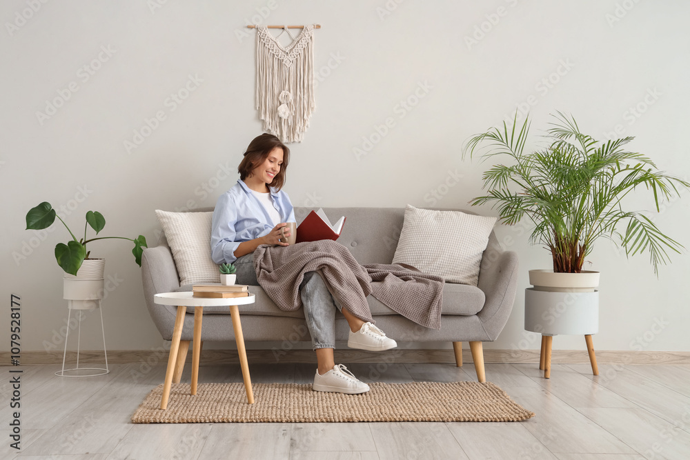 Fototapeta premium Pretty young woman with cup of tea reading book on sofa at home