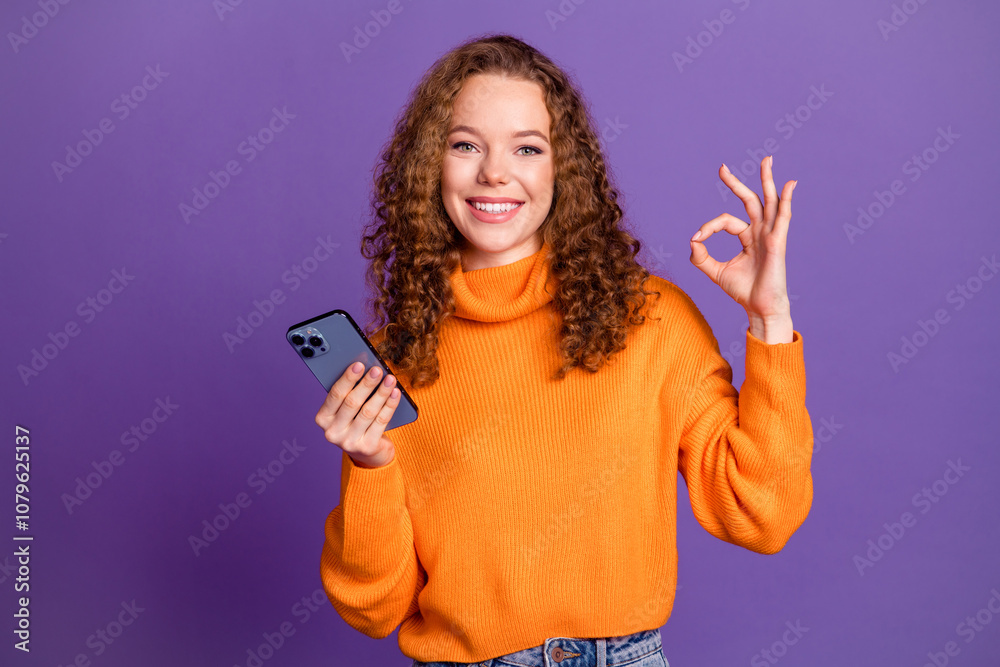 Portrait of pretty young girl smart phone okey symbol wear orange sweater isolated on purple color background Portrait of pretty young girl smart phone okey symbol wear orange sweater isolated on purple color background