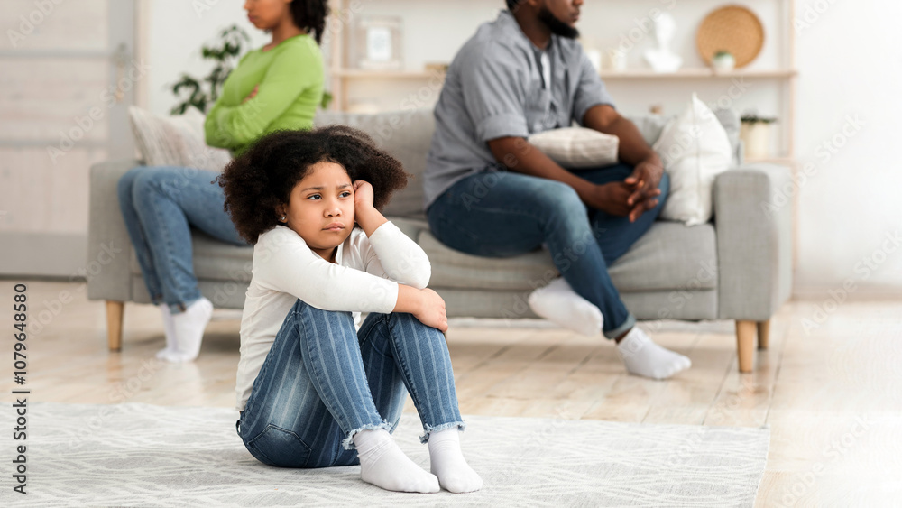 Family Quarrels Concept. Upset little black girl sitting on floor separately from parents after their argue, selective focus on kid Family Quarrels Concept. Upset little black girl sitting on floor separately from parents after their argue, selective focus on kid