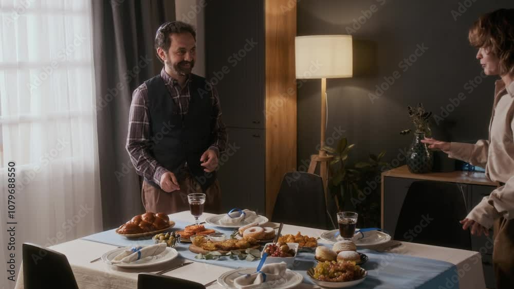 Zoom out shot of Jewish man in kippah serving challah on festive dinner table and talking with his wife while preparing for traditional Hanukkah celebration in cozy living room Zoom out shot of Jewish man in kippah serving challah on festive dinner table and talking with his wife while preparing for traditional Hanukkah celebration in cozy living room