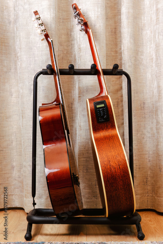 Two acoustic guitars are standing side-by-side on a black stand in a room. Musical studio backgrounds