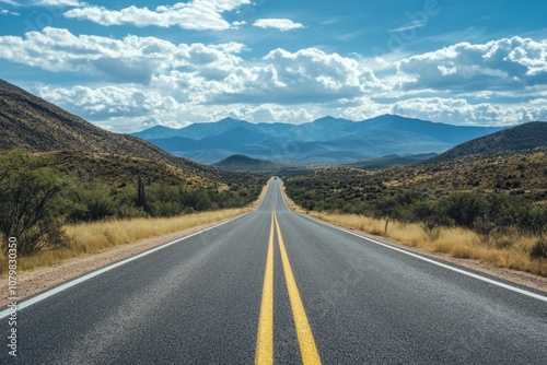 A long, straight road leads through a desert landscape towards a mountain range under a blue sky with clouds.