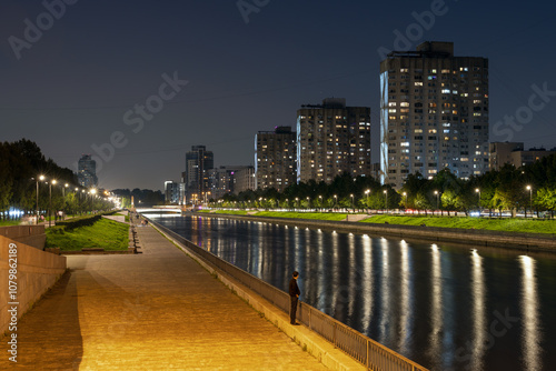 Smolenka River Embankment, Saint Petersburg, Russia. Evening cityscape. A lonely man stands on the embankment and looks at the river and multi-story residential buildings. Night city. photo