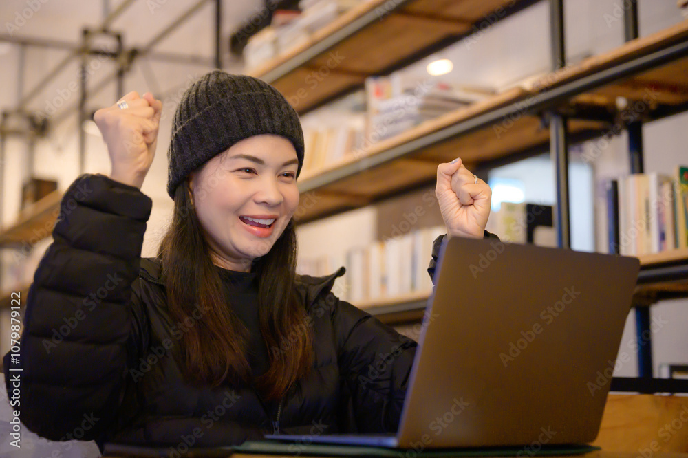 A young Asian woman celebrating success while working on a laptop in a cozy cafe, joyful freelancer achieving her goals, winter clothing. A young Asian woman celebrating success while working on a laptop in a cozy cafe, joyful freelancer achieving her goals, winter clothing.