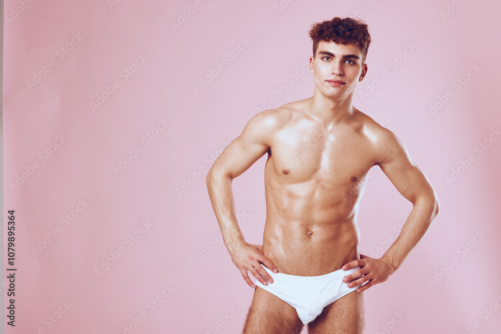 Fototapeta premium Confident young man posing in white swimwear against a soft pink background, showcasing fitness and body positivity. Fashion and lifestyle concept.