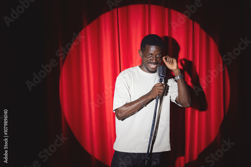 Smiling African American man speaking to microphone performing in comedy show open mike stand up on stage with red curtains. photo