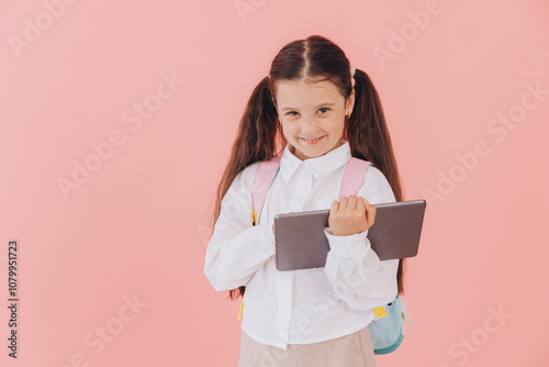 Schoolgirl with backpack holding tablet computer, isolated on pink studio background, looking at camera, smiling