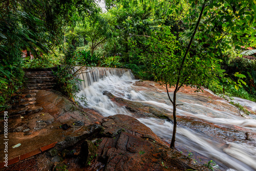 Natural background of various trees growing in the foothills of the mountain and natural waterfall flowing from high places amidst clear sky. The beauty of panoramic ecosystem.