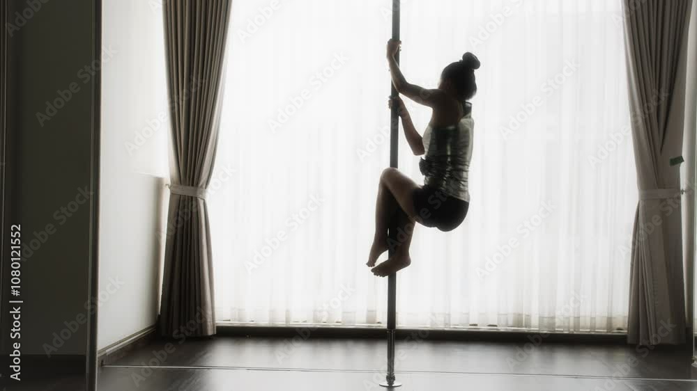 Long shot of silhouette of young woman performing pole dance in studio with natural light