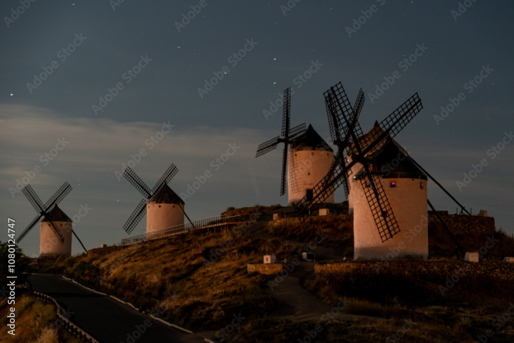 Naklejka premium A serene view of old windmills in Consuegra against a night sky, showcasing historic architecture illuminated by moonlight
