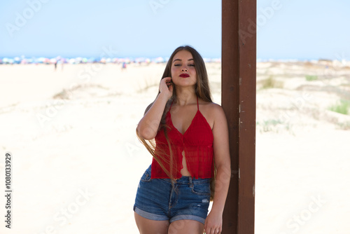 Young and beautiful Latin woman with long hair is leaning on a wooden poster on the beach. In the background the fine sand dunes with umbrellas and the sea and blue sky on the horizon.