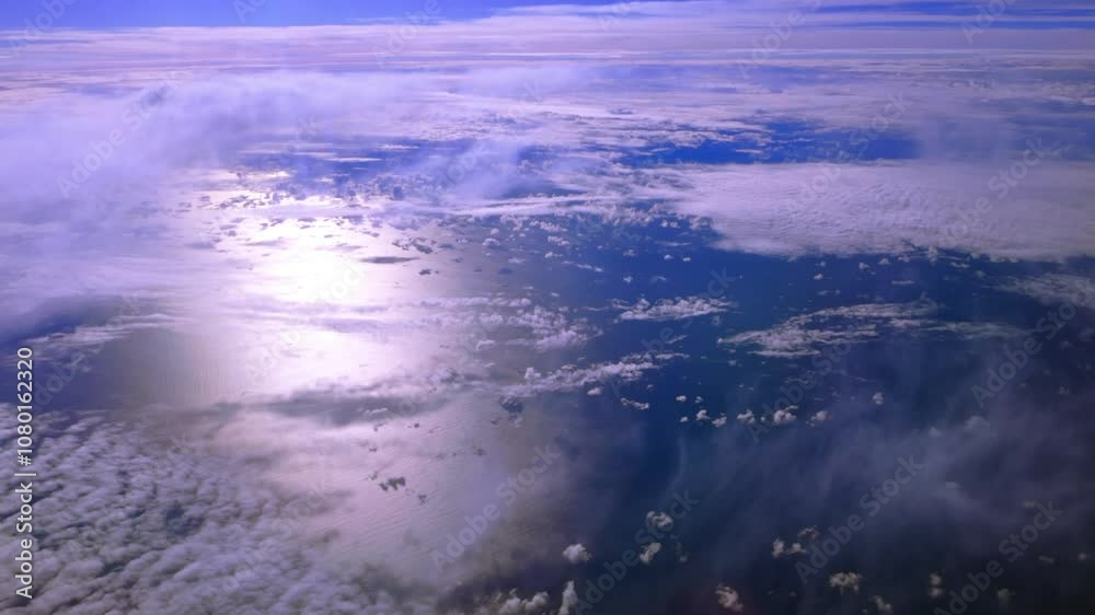 A high altitude aerial view over clouds and water on a sunny day, taken from the cabin of a passenger aircraft. The camera is stationary looking down out the port side window at the ocean and clouds. A high altitude aerial view over clouds and water on a sunny day, taken from the cabin of a passenger aircraft. The camera is stationary looking down out the port side window at the ocean and clouds.