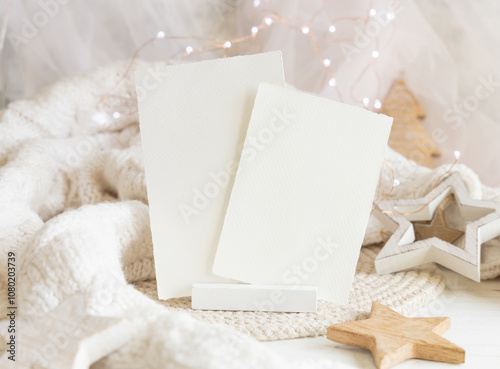 Paper cards near white sweater and wooden Christmas decor and lights, mockup