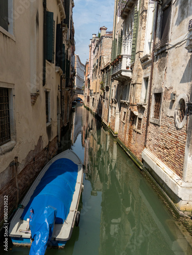 Wallpaper Mural Quiet Venetian canal with a small boat covered in blue tarp, surrounded by aged buildings with weathered facades. The scene captures the serene and timeless atmosphere of Venice, Italy. Torontodigital.ca