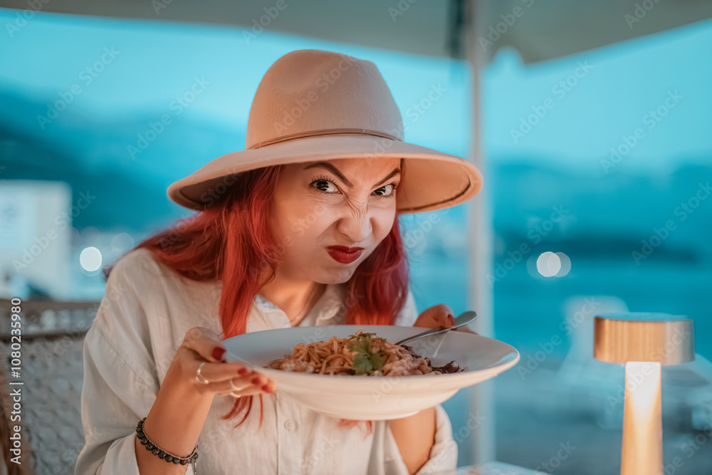 Redhead woman making a disgusted face while holding a plate of pasta in a restaurant by the sea