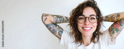 Portrait of a beautiful young tattooed female model looking at camera holding both her hands behind her head, isolated on grey background. Wearing white t-shirt.