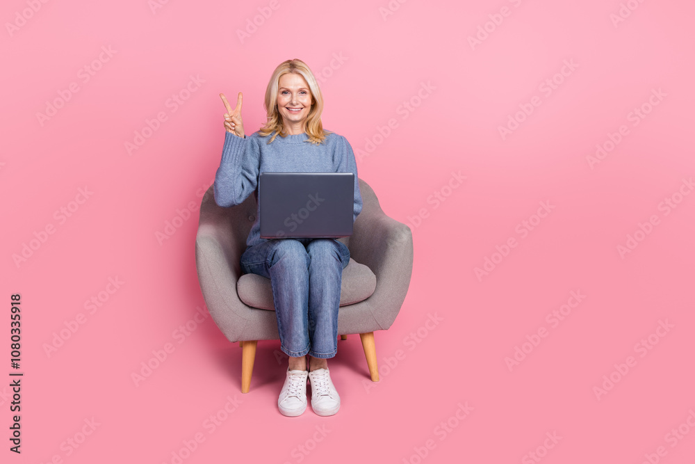 Full body photo of attractive senior woman sit armchair netbook v-sign dressed blue knitted clothes isolated on pink color background Full body photo of attractive senior woman sit armchair netbook v-sign dressed blue knitted clothes isolated on pink color background