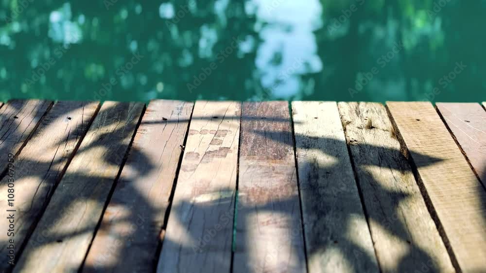 Space on a vintage wooden plank dock or table with the shade of a tree on the floor, the background of the turquoise pond under sunlight in the morning.