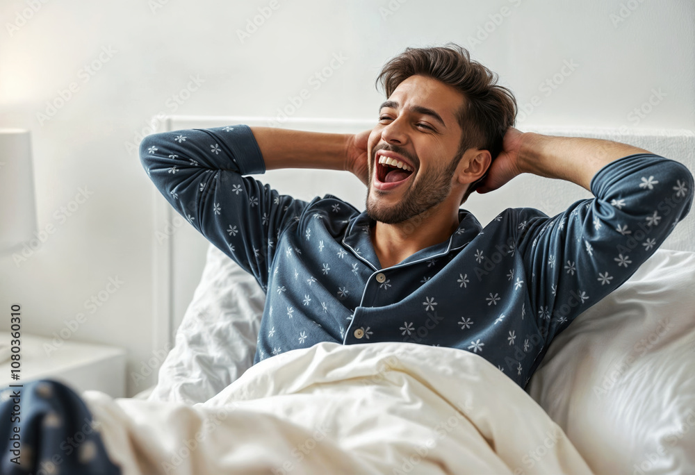 Cheerful young Indian man in floral pajamas laughing while relaxing in bed, wearing wireless earbuds and enjoying lazy morning at home