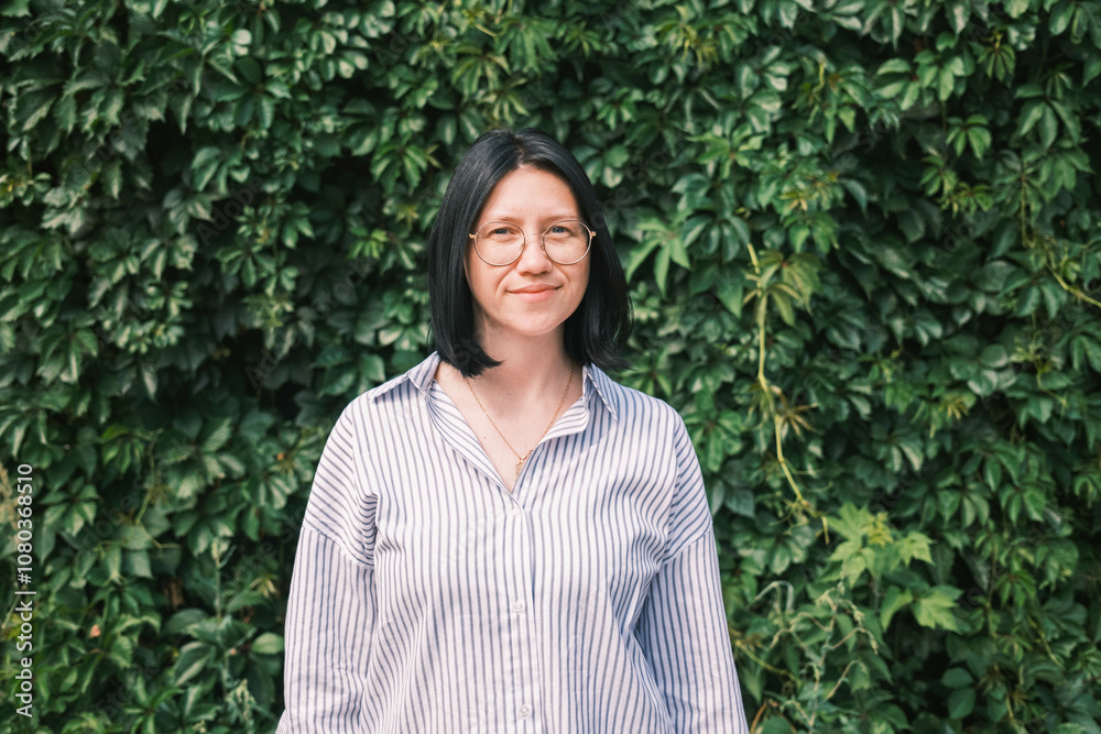 A beautiful young woman with black bob hair, wearing glasses and a light striped summer shirt, stands against the background of green grape leaves. A beautiful young woman with black bob hair, wearing glasses and a light striped summer shirt, stands against the background of green grape leaves.