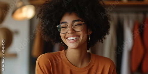 joyful woman with curly hair and glasses smiles brightly, showcasing her happiness in cozy setting. Her warm expression radiates positivity and confidence.