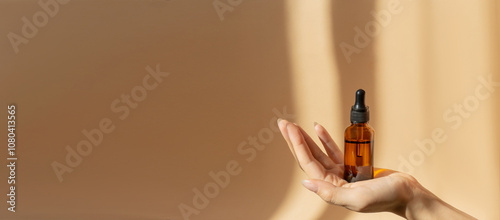 Bottle of serum in women's hands. Glass bottle with dropper cap on a beige banner with a copy space. Unmarked Amber container in sunlight