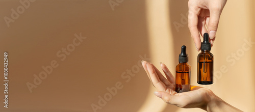Women's hands hold bottles of amber glass with cosmetic serum on brown background. Mockup of container with dropper lid with collagen in rays of sunlight. Copy space,banner