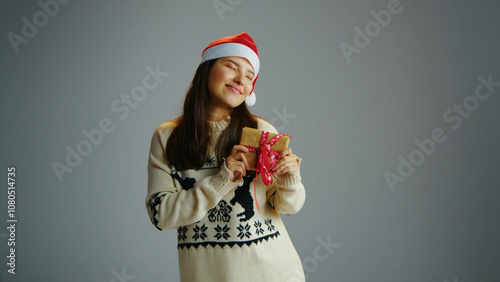 Fun cute woman hold small Christmas gift on gray isolated studio background. Joyful girl give new year eve present. Person wear red Santa Claus hat receive packaging box. Winter holiday xmas surprise.