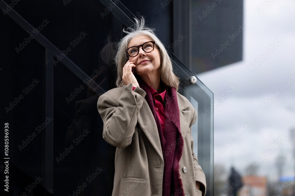 Fototapeta premium Elderly businesswoman talking on the phone outside her office building during a cloudy day