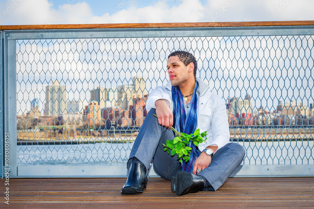 A man sits on a wooden deck with a city skyline in the background, holding a bunch of green leaves. He's dressed in a white jacket and blue scarf, looking thoughtfully into the distance.