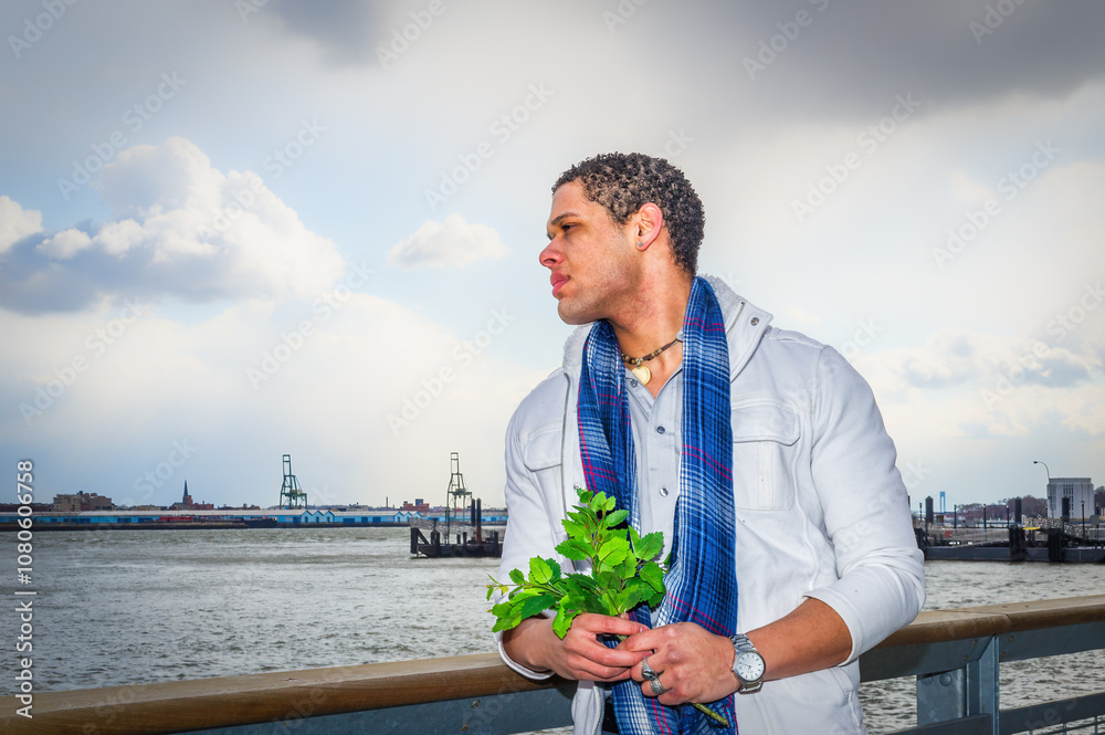 A man wearing a blue scarf stands by a waterfront, holding a bunch of green leaves. The background features industrial structures and a cloudy sky.