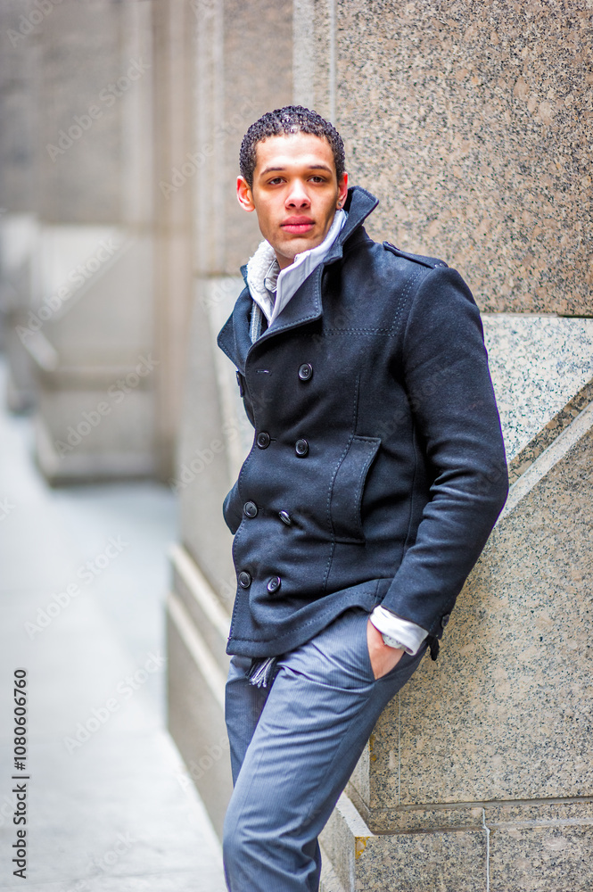 A stylishly dressed man leans against a stone wall in an urban setting. He wears a dark double-breasted coat with a popped collar and light gray trousers.