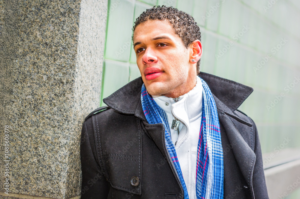 A person with short curly hair is leaning against a textured wall, wearing a dark coat and a blue plaid scarf. The background features a green, glass-tiled surface, adding a modern urban feel.