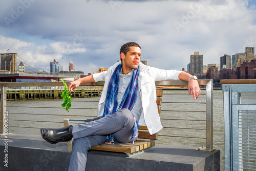 A man dressed stylishly in a white jacket and a blue scarf is seated on a bench by the waterfront, with greenery in hand. The city skyline and a prominent bridge are visible in the background. photo