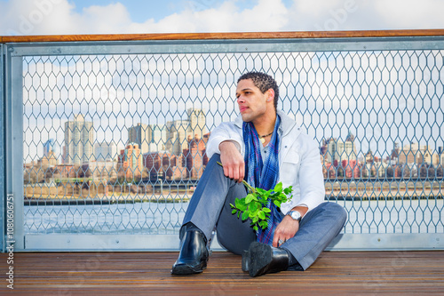 Obraz na płótnie A man sits on a wooden deck with a city skyline in the background, holding a bunch of green leaves. He's dressed in a white jacket and blue scarf, looking thoughtfully into the distance. w salonie