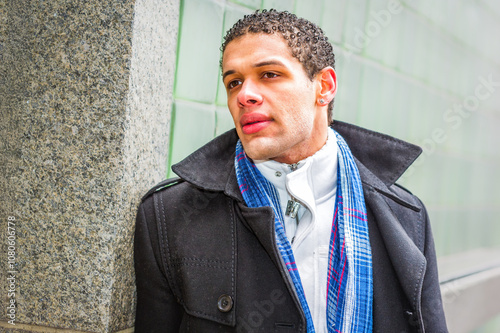 Obraz na płótnie A person with short curly hair is leaning against a textured wall, wearing a dark coat and a blue plaid scarf. The background features a green, glass-tiled surface, adding a modern urban feel. w salonie