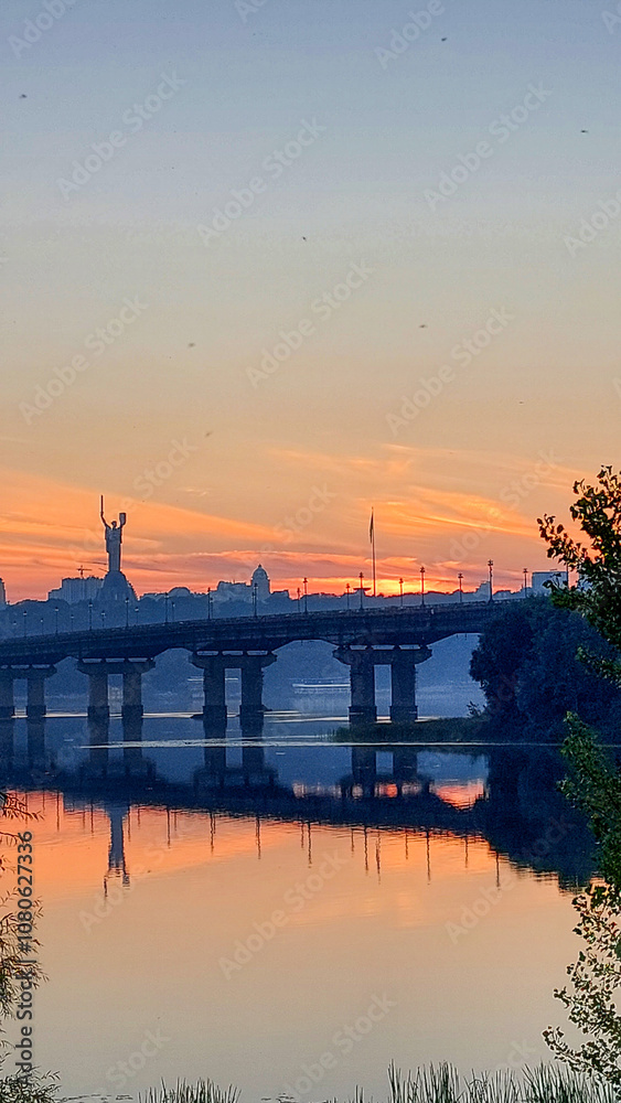 Captivating sunset view in Kyiv, Ukraine, with the famous Motherland Monument and bridge reflected in the calm waters of the Dnipro River Captivating sunset view in Kyiv, Ukraine, with the famous Motherland Monument and bridge reflected in the calm waters of the Dnipro River