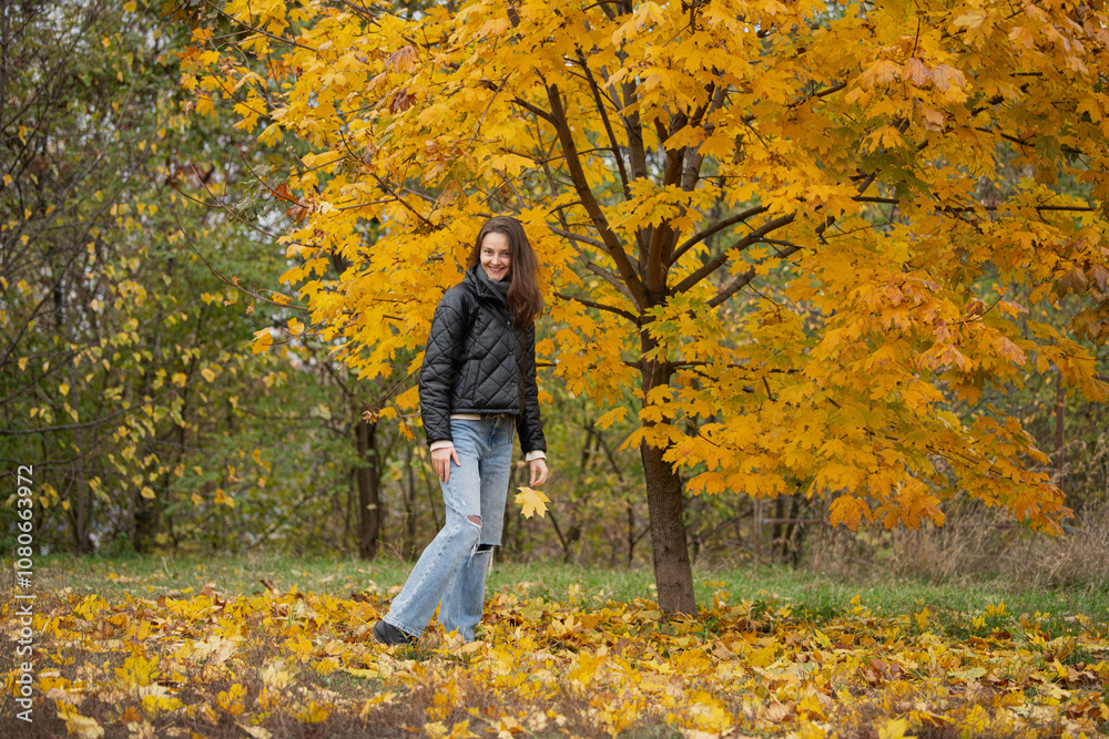 A young woman strolls through a park blanketed in yellow and orange leaves, surrounded by the warm colors of autumn, enjoying the tranquil atmosphere.