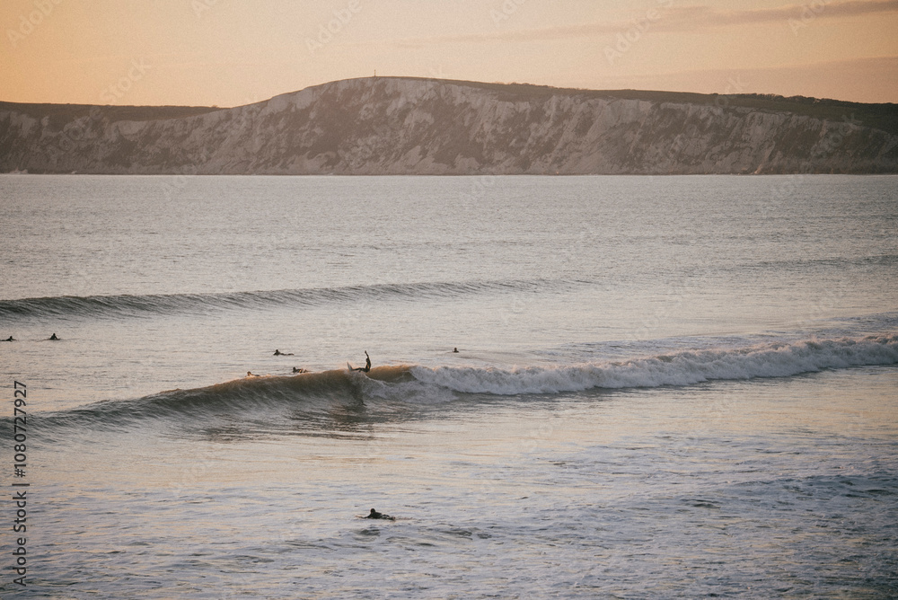 Fototapeta premium Person surfing on the waves of the sea near the coastal island and people swimming in the sea in evening