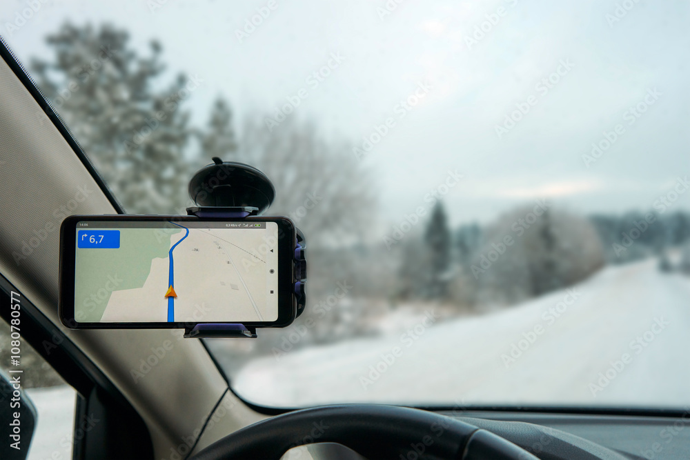 Navigation application on a smartphone attached to the windshield of the car inside the cabin. Snowflakes on the windshield, snow-covered road and forest through the glass. Navigation application on a smartphone attached to the windshield of the car inside the cabin. Snowflakes on the windshield, snow-covered road and forest through the glass.