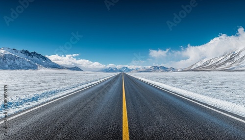 A long, empty road stretches through a snowy landscape under a clear blue sky, surrounded by distant mountains.