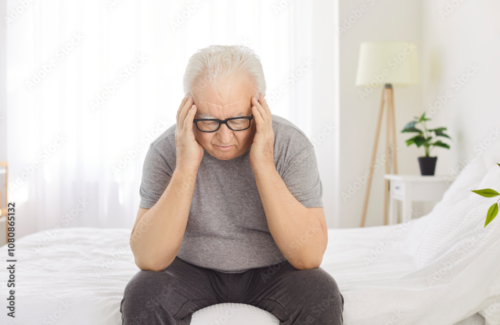 Sick elderly man is suffering from a headache or migraine while sitting on bed at home. Health problems, with the man feeling stressed, sad, and in pain, indicating his discomfort. Sick elderly man is suffering from a headache or migraine while sitting on bed at home. Health problems, with the man feeling stressed, sad, and in pain, indicating his discomfort.