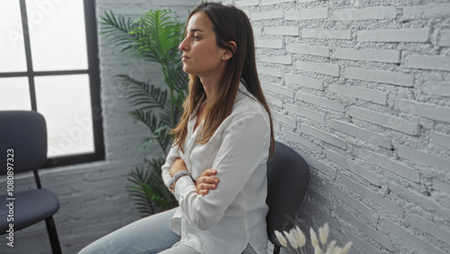 Young woman with crossed arms sitting ina corridor with a brick wall and plants background indoors photo