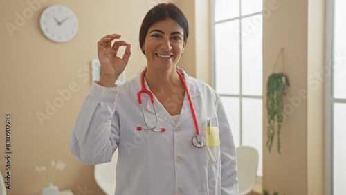 A young, beautiful hispanic woman doctor with brunette hair stands in a clinic room, smiling and making an okay gesture with her hand. photo