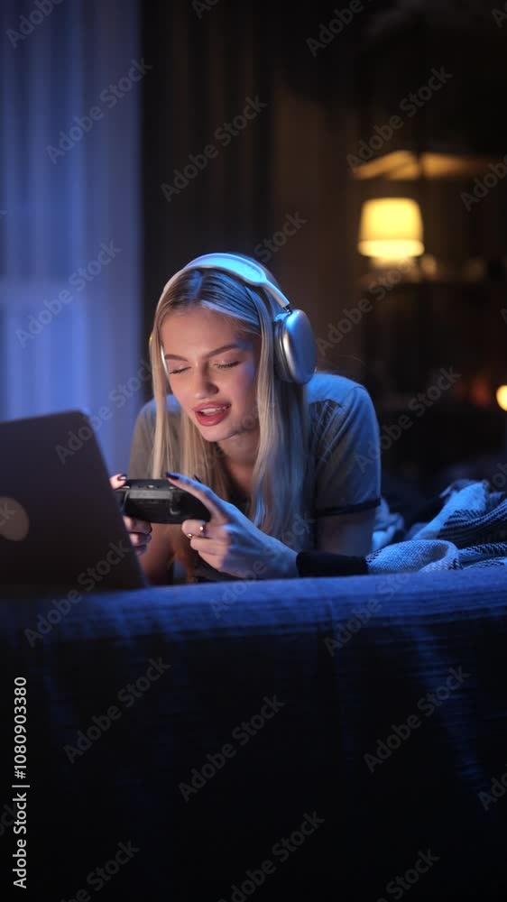 A Young Gamer is Enjoying a Late Night Gaming Session in the Comfort of Their Home. Cheerful Blonde Woman Wearing Headphones and Using her Laptop. Vertical Shot A Young Gamer is Enjoying a Late Night Gaming Session in the Comfort of Their Home. Cheerful Blonde Woman Wearing Headphones and Using her Laptop. Vertical Shot