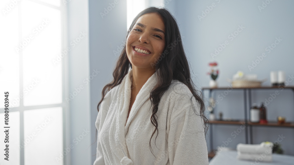 Beautiful woman smiling in a spa wearing a white robe with a wellness setting in the background showing towels and candles on shelves Beautiful woman smiling in a spa wearing a white robe with a wellness setting in the background showing towels and candles on shelves