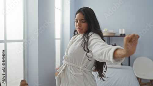 Young brunette woman in a white robe stretches in a serene spa room with soft natural light, evoking wellness and relaxation.